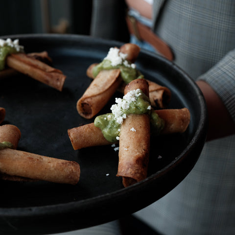 A close-up of a black serving tray holding several rolled appetizers topped with green sauce and white crumbles, with part of a person in a gray plaid blazer holding the tray.